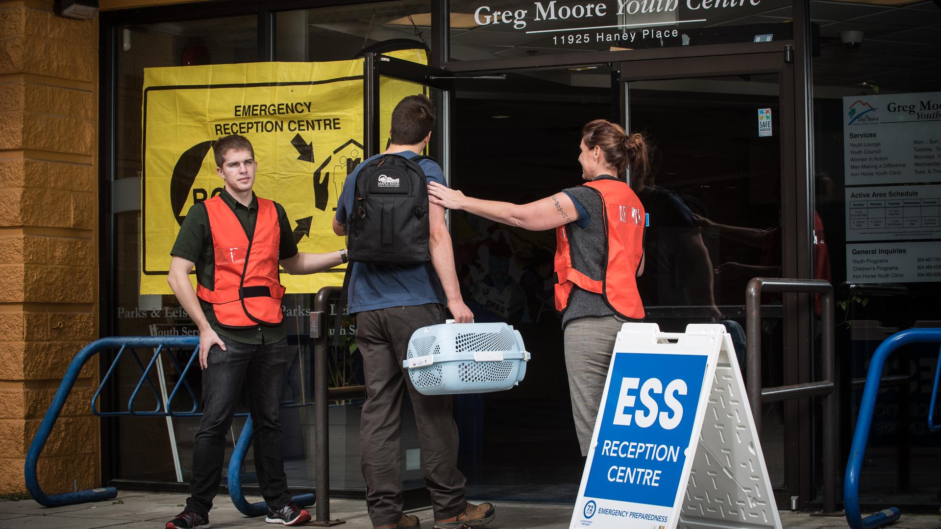 A pair of workers in high-visibility vests welcome a man carrying a toolbox to an Emergency Support Services reception centre.