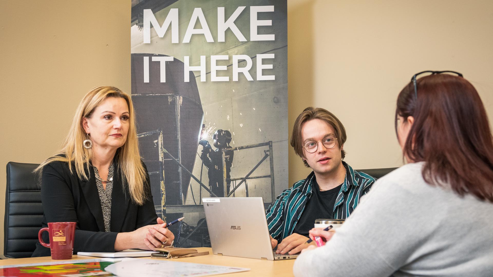 A group of people have a discussion in front of a sign that says "Make it here".