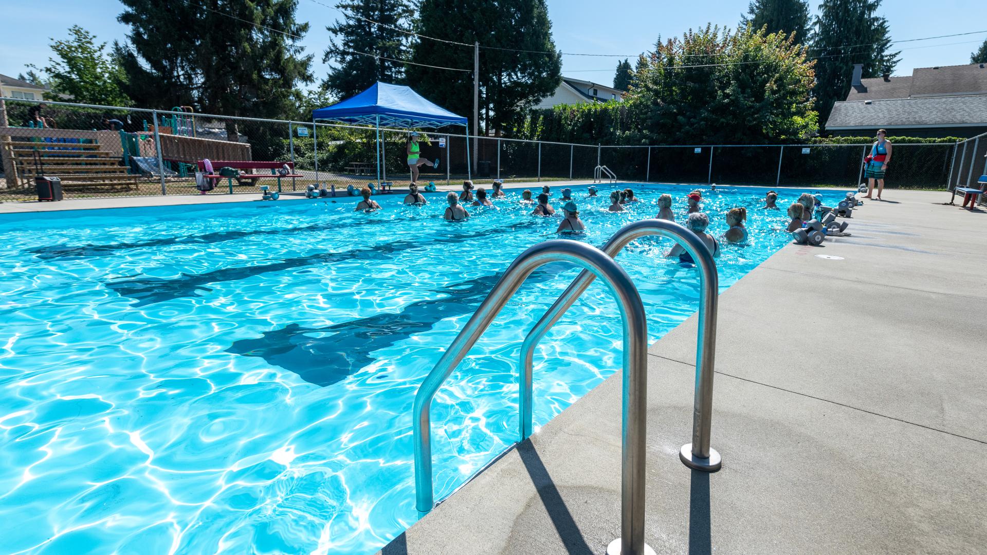 The assistance railing at the Hammond outdoor pool gleams in the sunlight while patrons enjoy the waters at the far end.