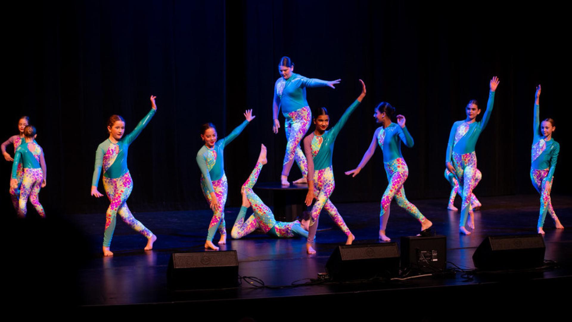 A group of young girls in matching blue leotards perform a dance routine on a black stage and background.