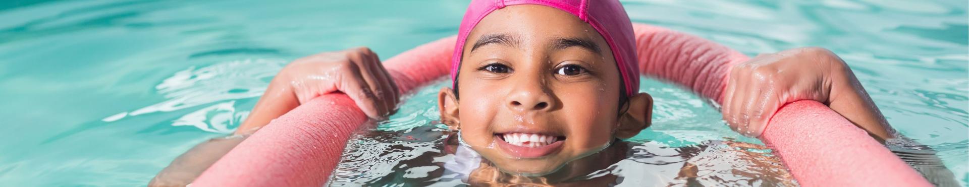 Child floating in pool wearing pink cap and holding pink floatation device