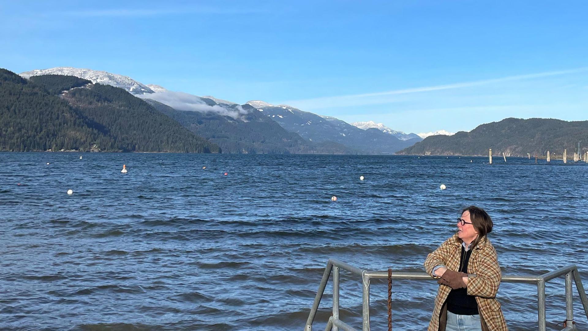 Panorama of mountains. On the foreground, Colleen Brown is leaning against railing. Colleen Brown is waring a brown coat with gloves. Behind Colleen Brown there is a large body of water. 