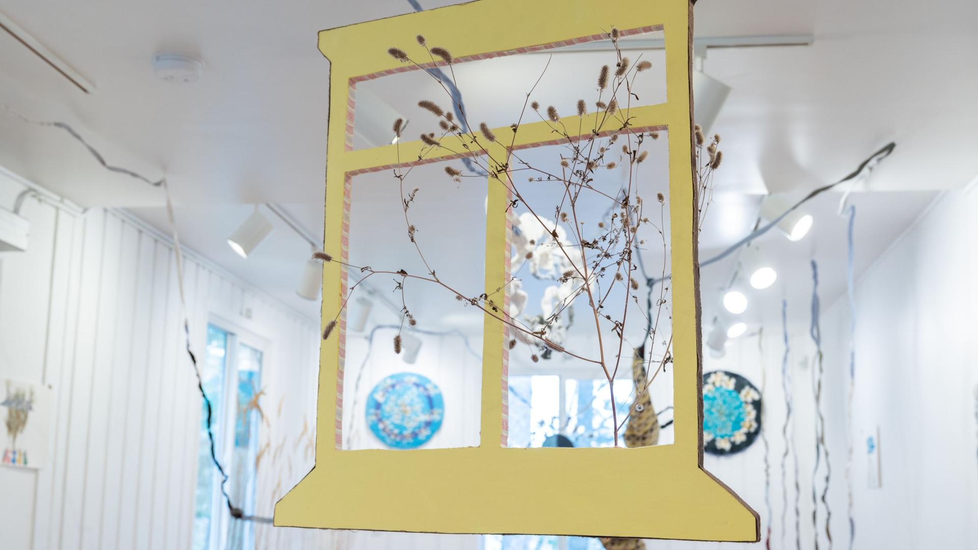 Piece of art by Colleen Brown. There is a yellow wooden window hanging in a room with various other pieces of art. 