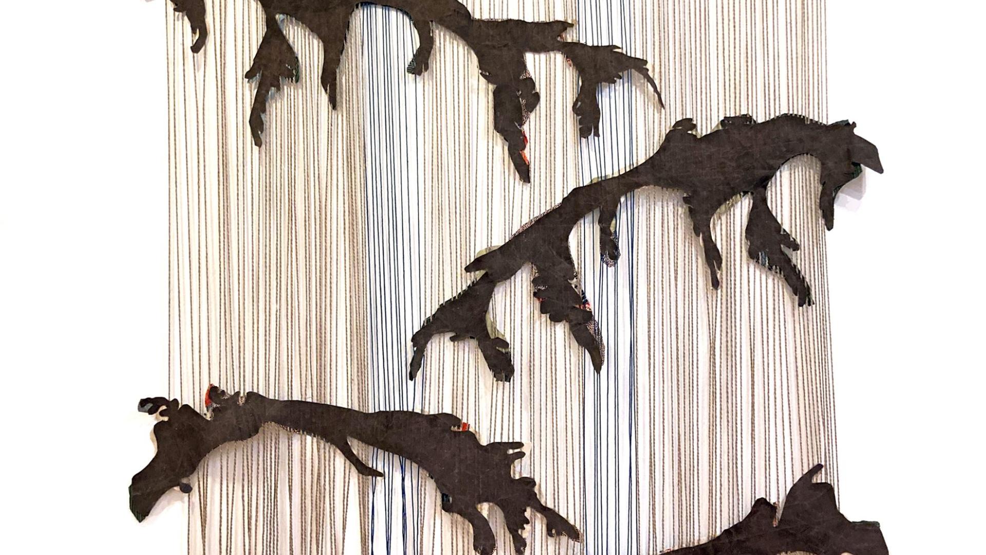 Piece of art by Colleen Brown. There is what looks like a tapestry hanging from a wall. Interwoven between the gapped threads, there are silhouettes of tree branches. The art piece is hanging from a being wall.