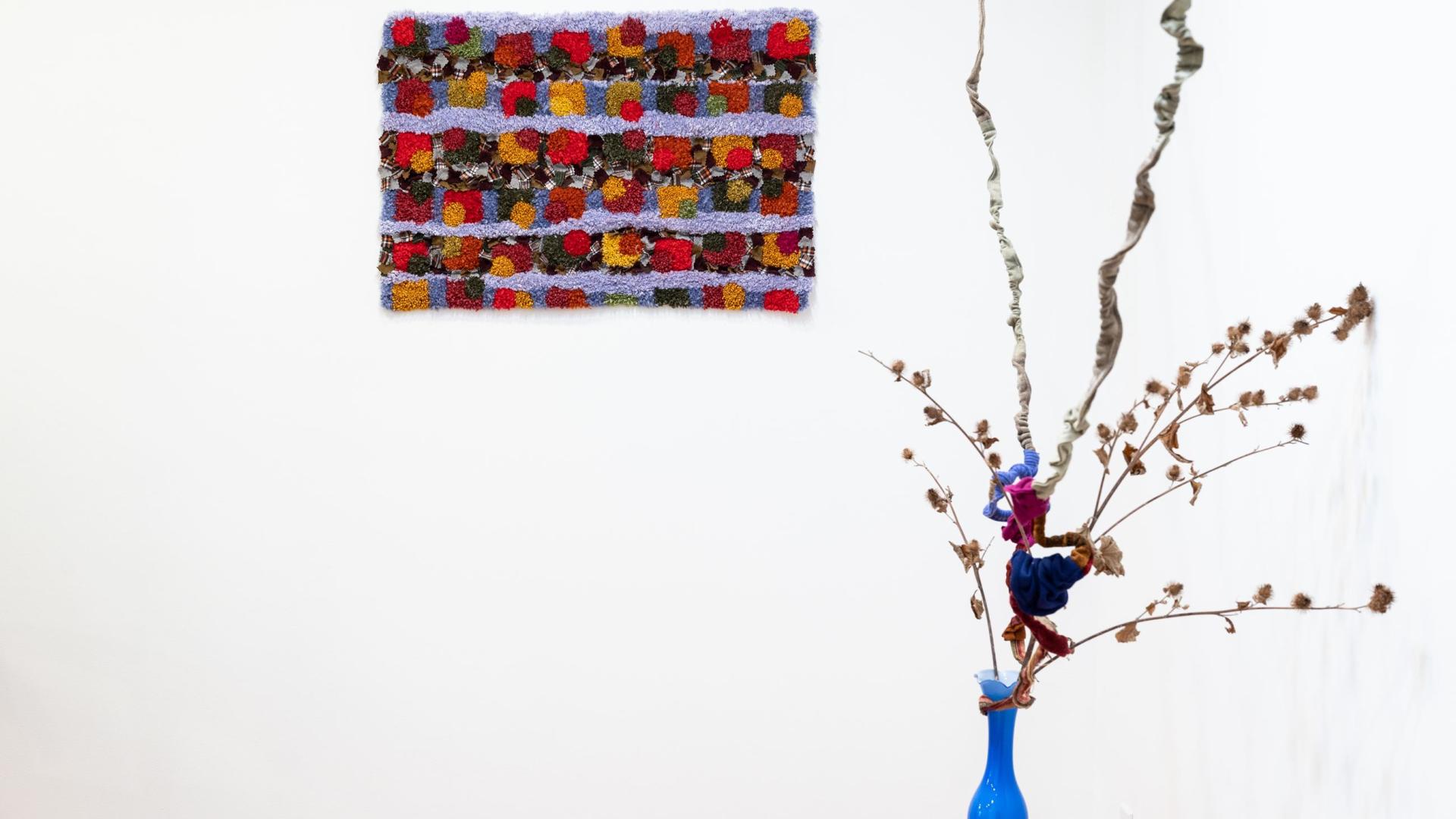 Artwork by Colleen Brown. There is a vase with flowers in it and a small rectangular piece of art made up of different coloured textiles. The works of art are displayed in front of a beige wall.