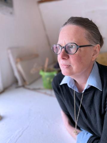 Portrait image of Colleen Brown. There is a person with glasses, necklace, and sweater over collared shirt with their gaze facing the left. They have their arms perched on top of a table.
