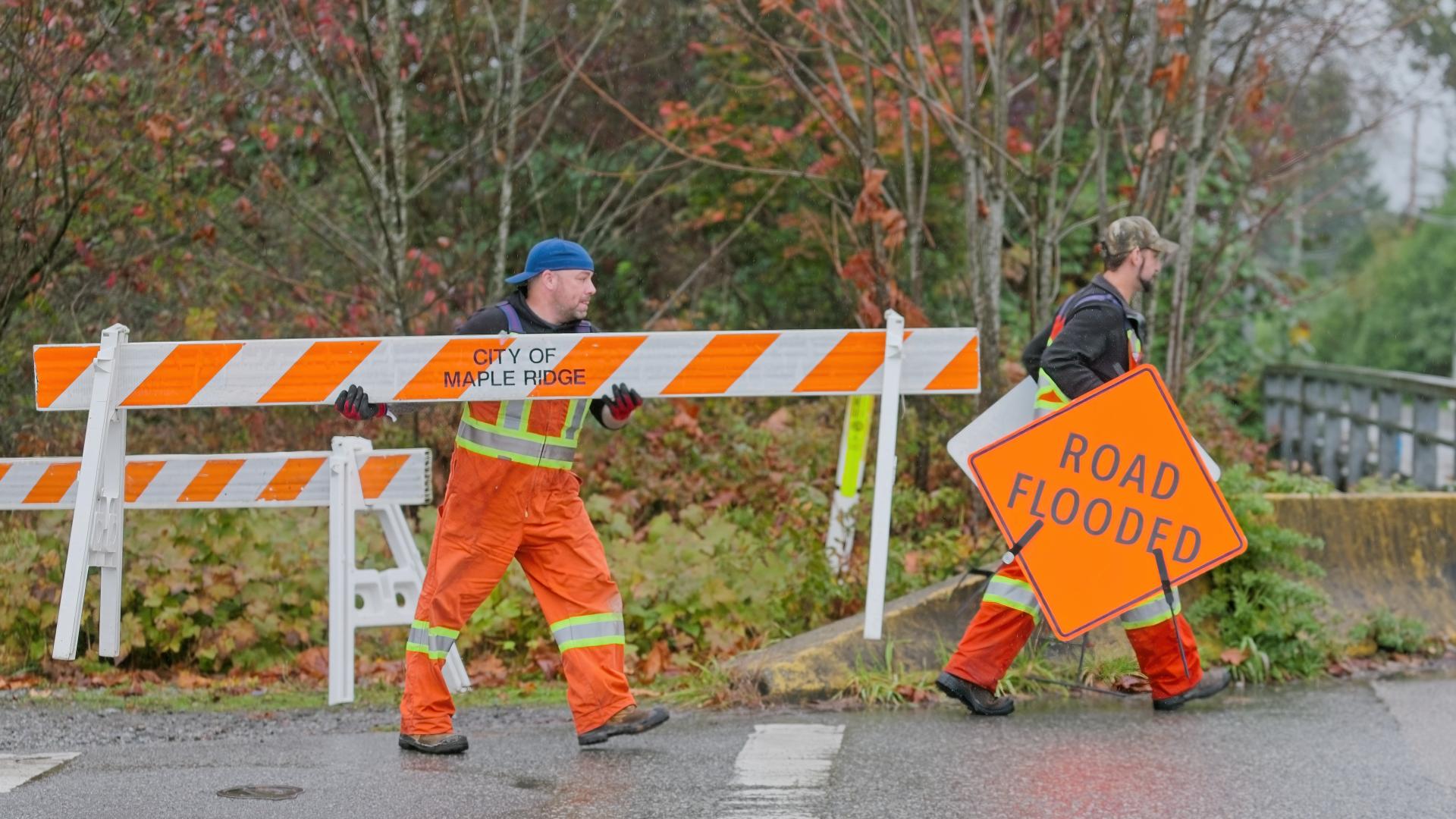 One man carries a sign saying "Road Flooded" while another follows carrying a wooden barricade stating "City of Maple Ridge".