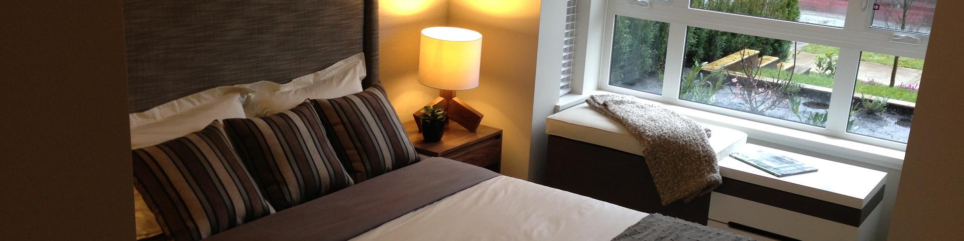 A well-made bed in warm browns and creams with a lamp sits next to a cool window, showing a rainy day outside.