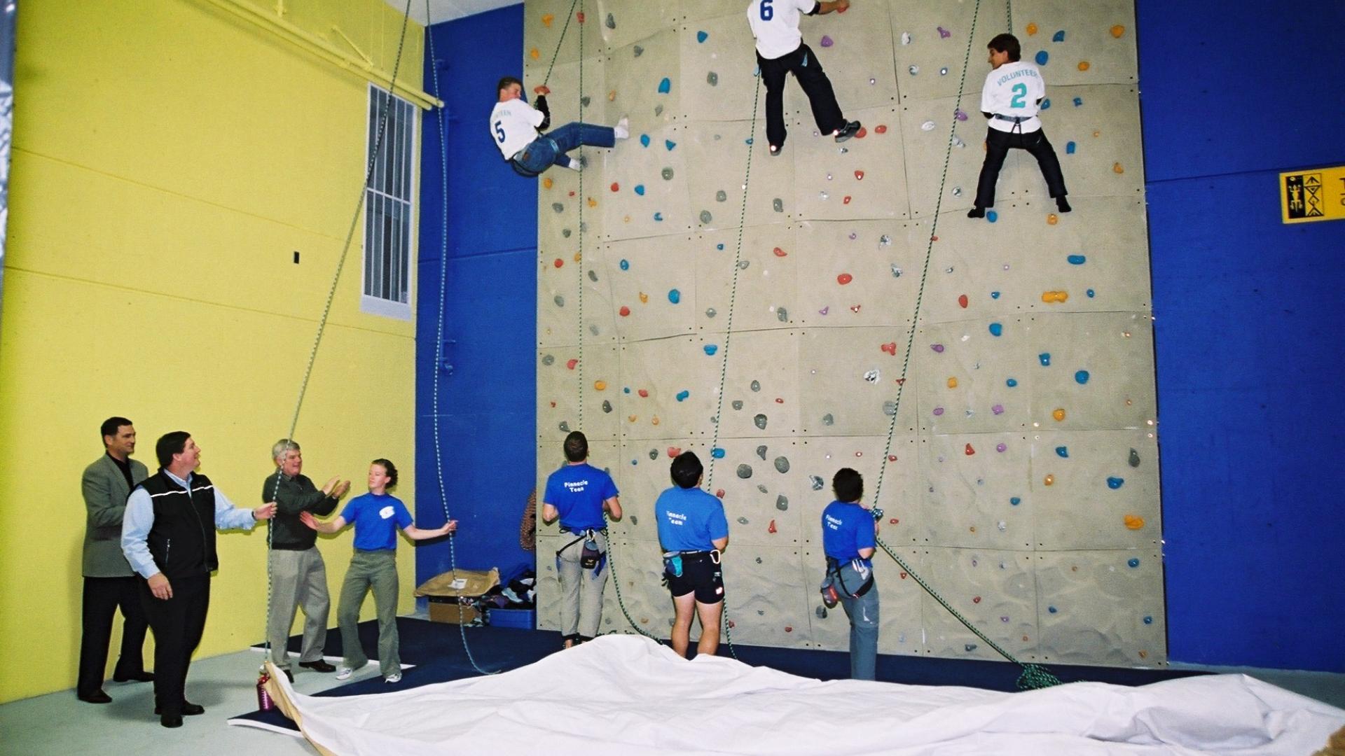 Three youths climb an indoor rock wall while staff belay them, and others watch.