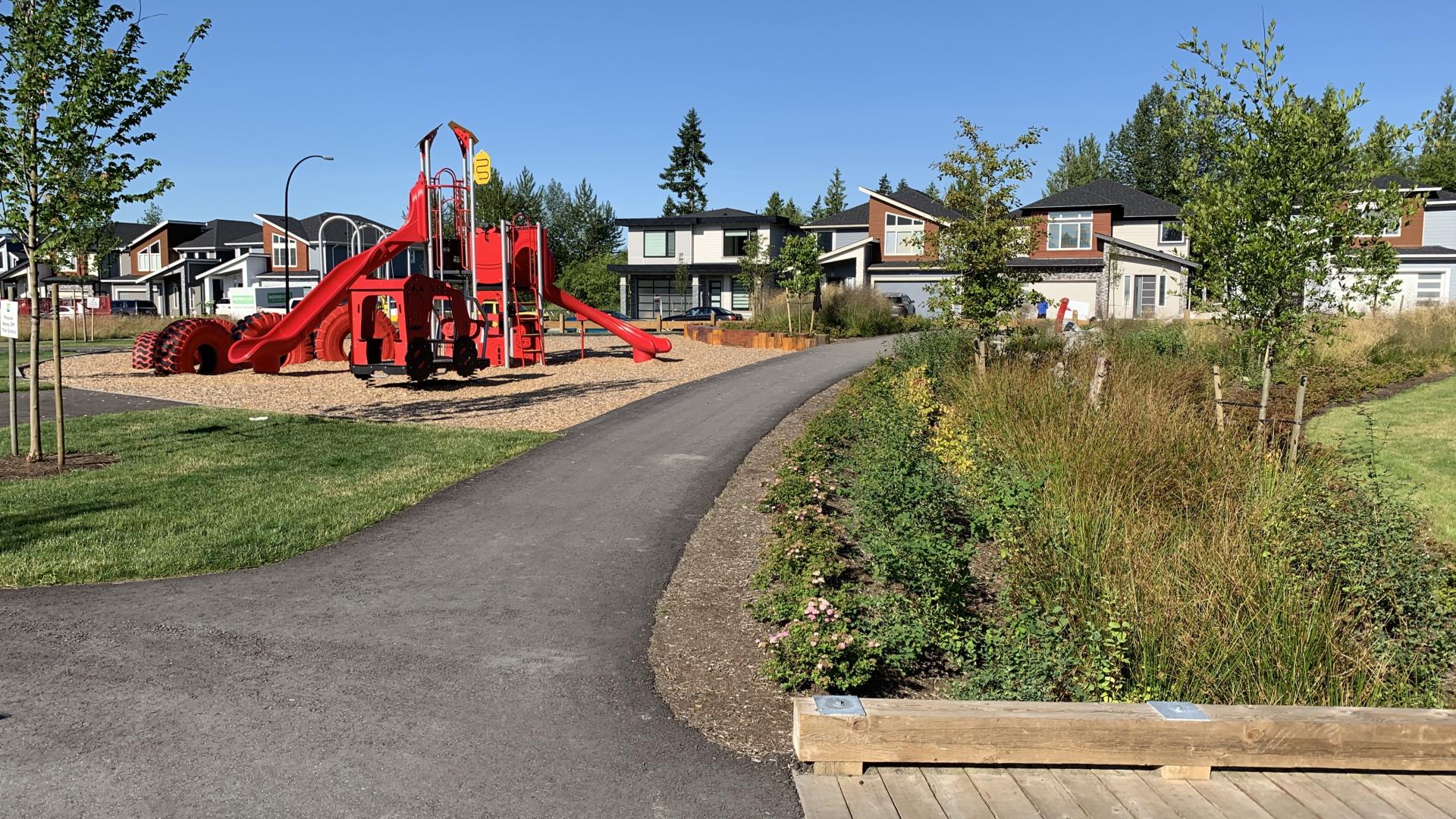 Paved path through neighbourhood park with red playground in the distance and wooden bridge on the right