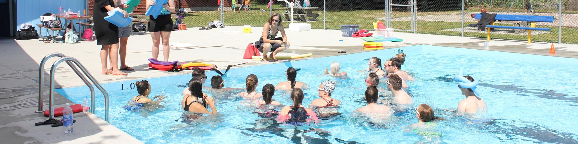 An instructor squats next to the Hammond Outdoor Pool, which is full of people, to teach.