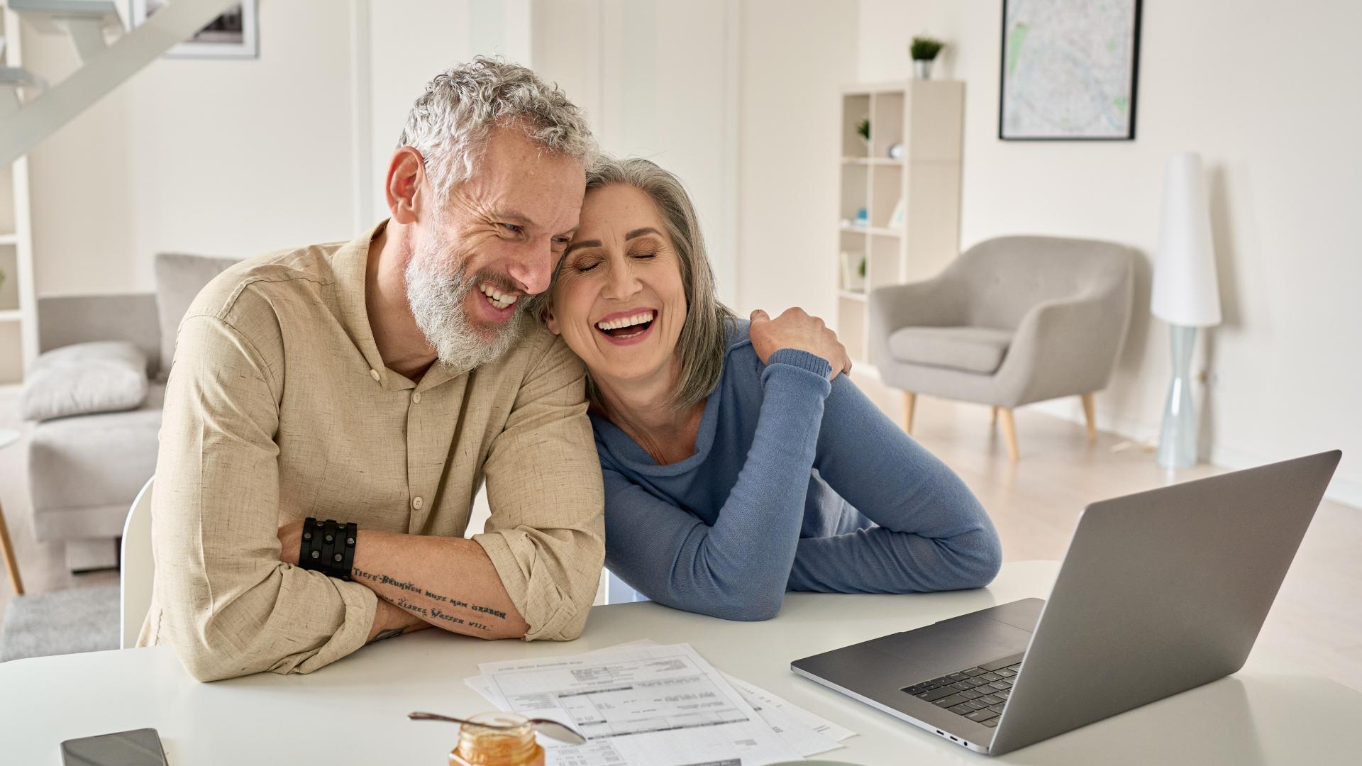 A man and a woman laugh together while looking at a computer on their kitchen table.