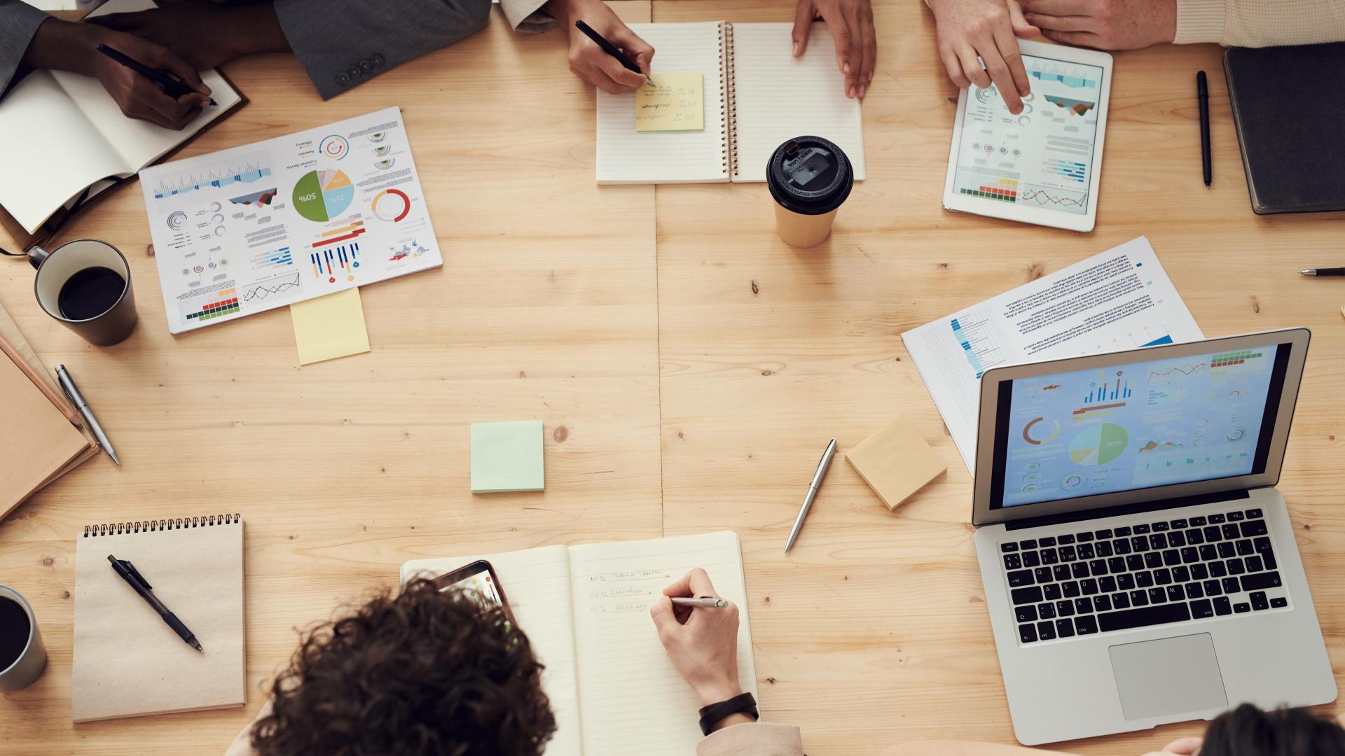 A group of people sit around a table, working collaboratively with computers, pens and paper.
