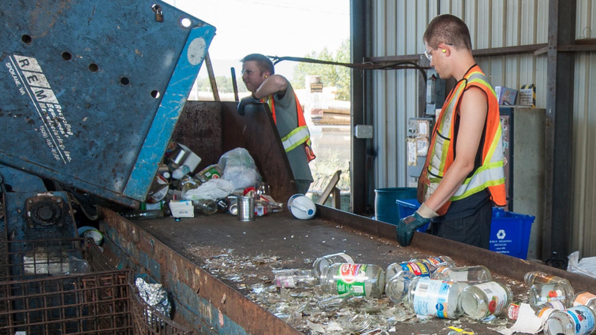 A man wearing safety gear draws glass out of a machine and onto a conveyer belt while another man wearing similar gear supervises.