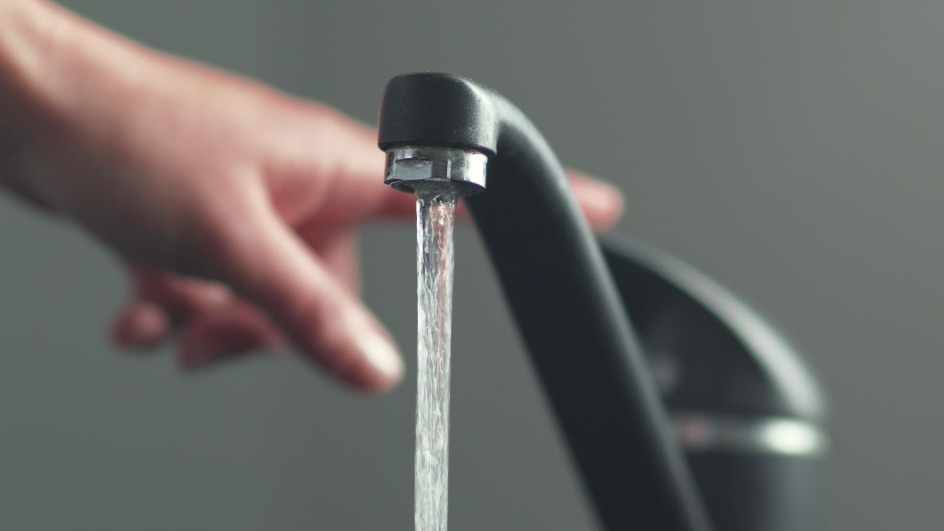 A hand turns on a tap, causing water to flow out of a faucet.