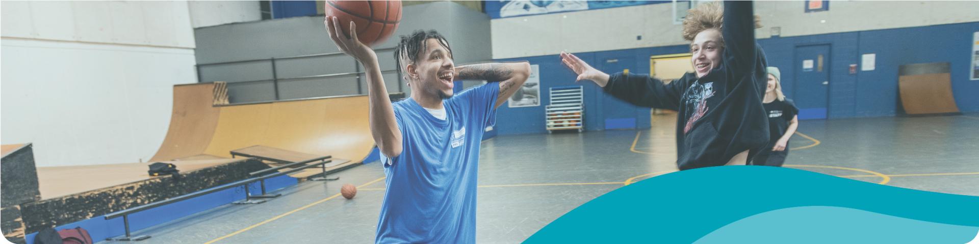 A young man reaches out to shoot a basketball pass his blocking opponent in the game.
