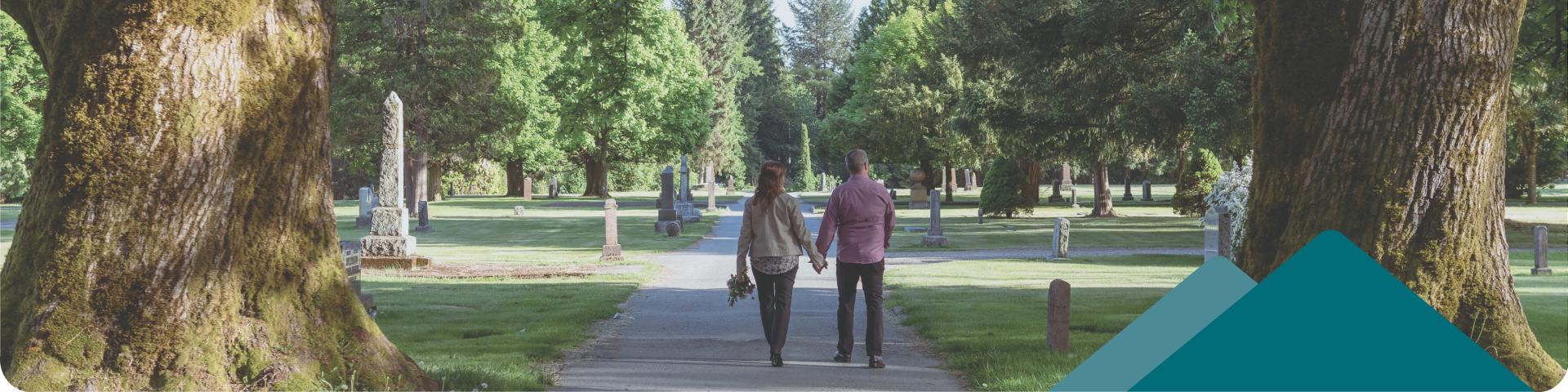 Two people walking holding hands through a cemetery with large trees.