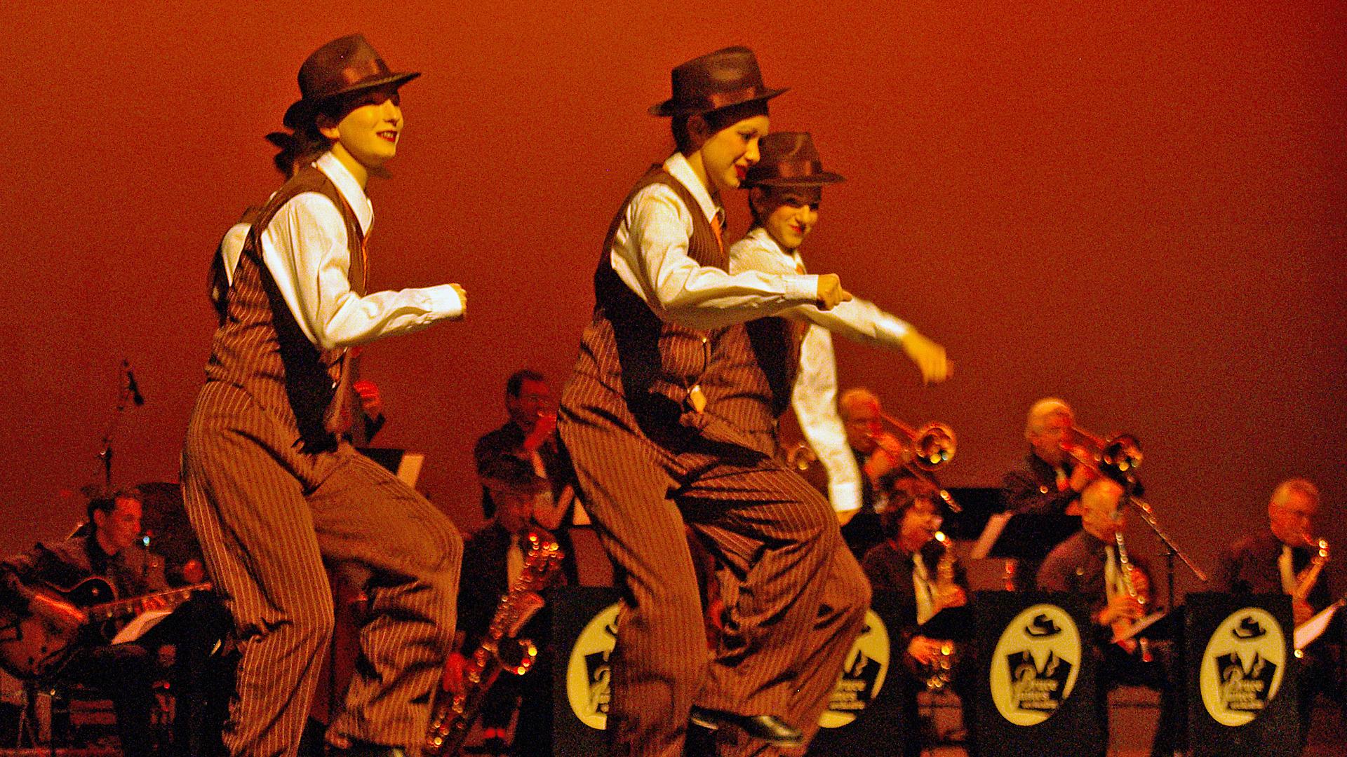 A group of female performers tap dance on a red stage with the accompaniment of a live band.