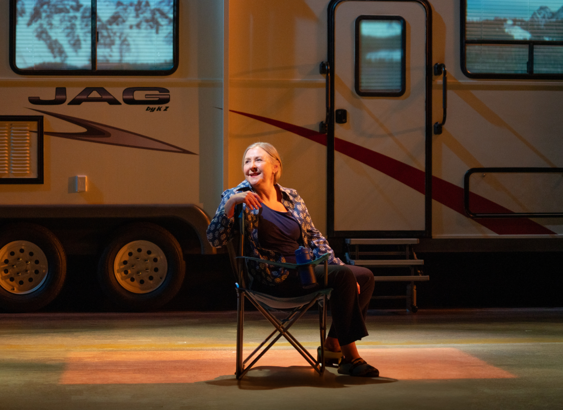 A female performer sits onstage in a camping chair outside a recreational vehicle.
